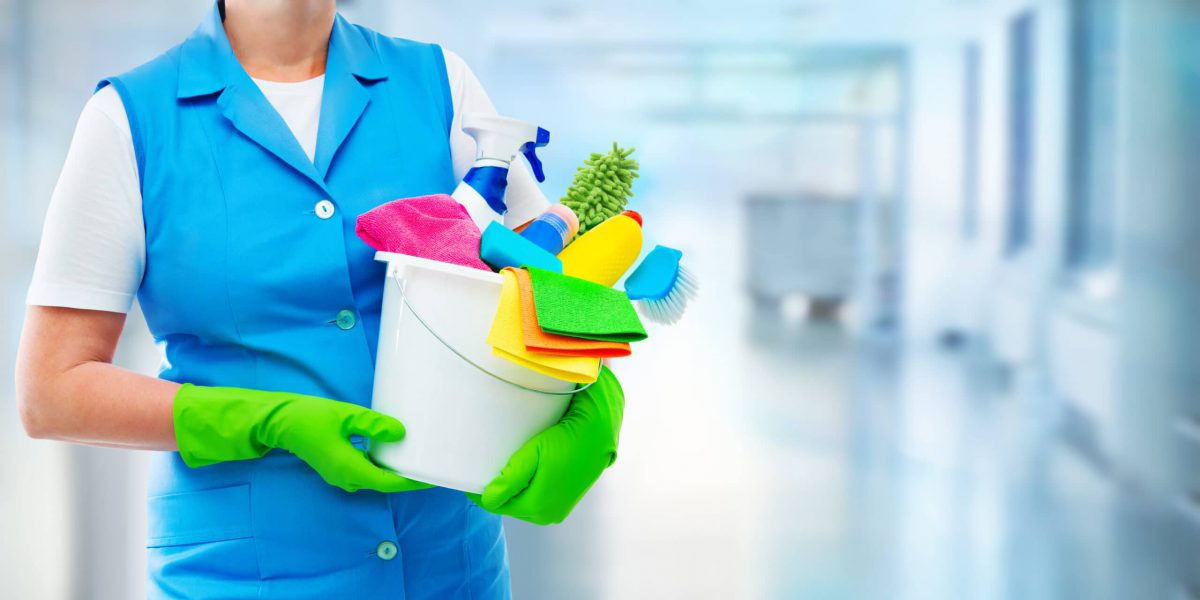 Female housekeeper while cleaning office. Woman wearing protective gloves and holding bucket full of cleaning supplies on blurred background