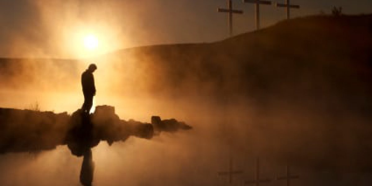 Dramatic religious photo illustration of Good Friday and Easter Sunday Morning reflecting a prayerful moment of silence with a silhoutted person bowing his head, a warm sunrise rises over a foggy lake, and three crosses on a hill reflected in the water as well.